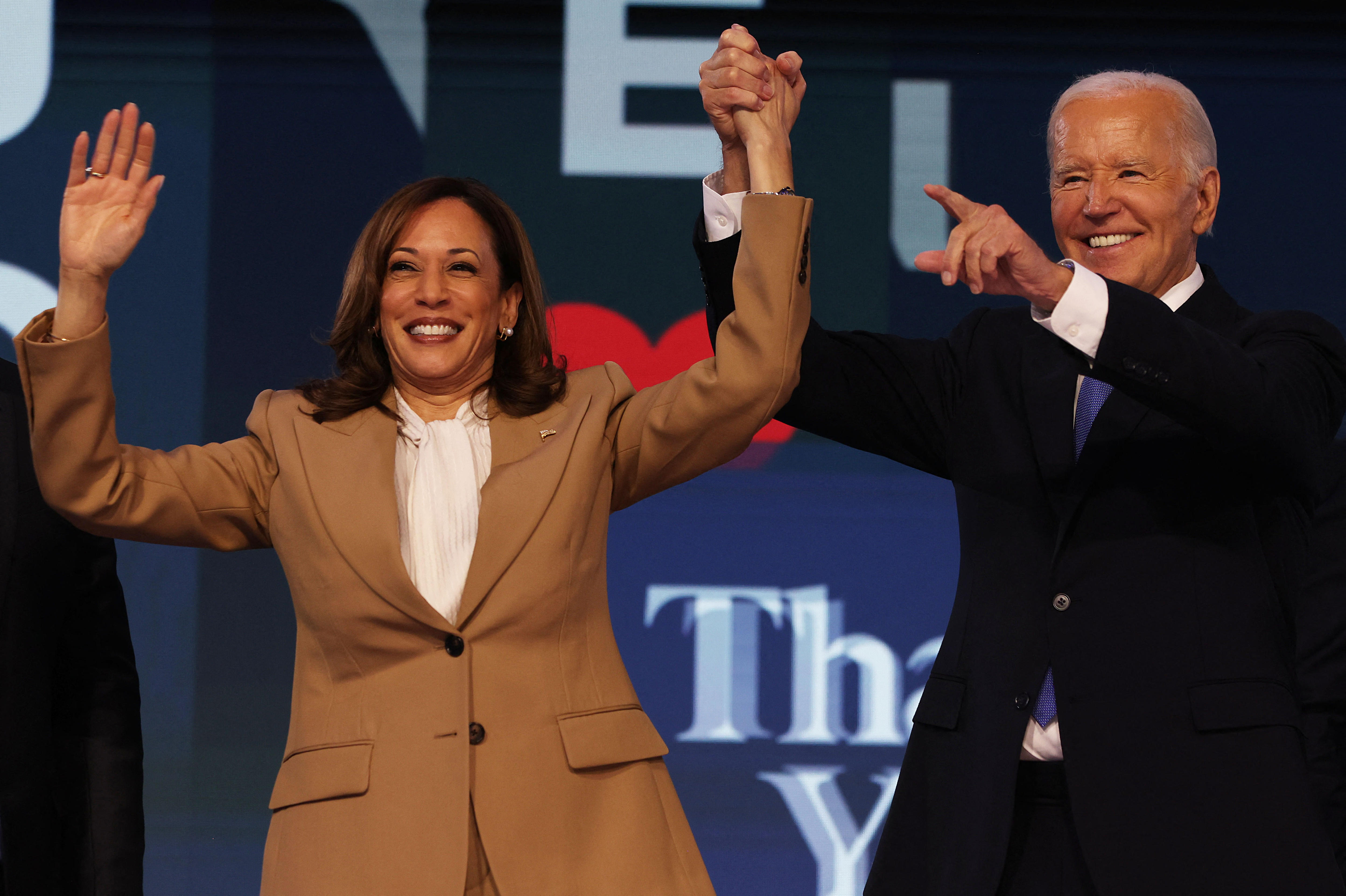 Joe Biden smiles and points as he holds aloft Kamala Harris's arm in celebration on a blue stage.