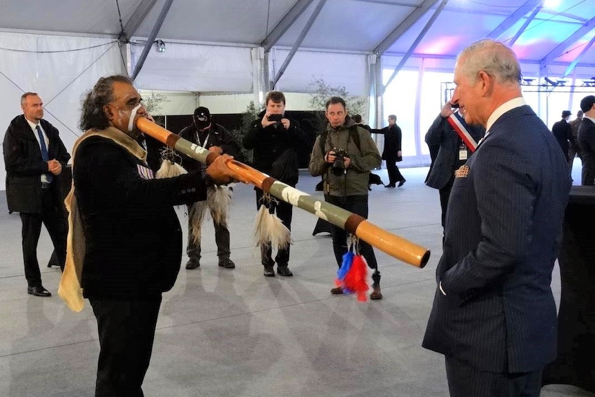 An Aboriginal man in a suit with a painted face plays the didgeridoo directly towards the stomach of a smiling Prince Charles.