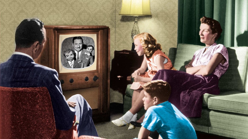 An advertisement showing a family watching a television set. The screen shows a moustached man holding ventriloquist dolls.