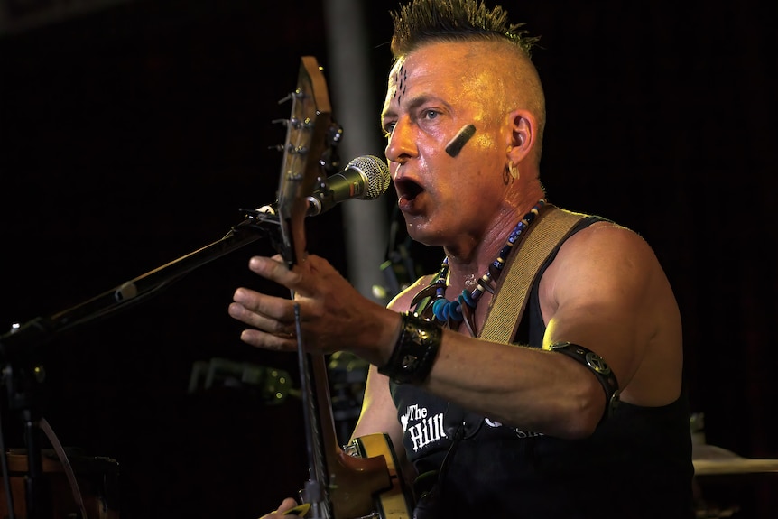 A man with a mohawk sings into a mic while playing a banjo.