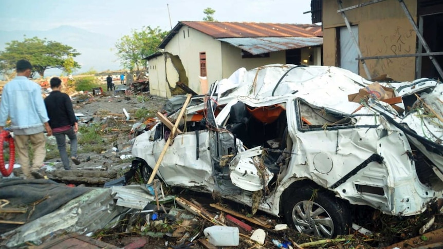 The wreckage of a mangled white car sits on a layer of debris, including mud, wood and rubbish, as two men walk past