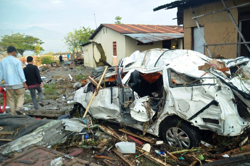 The wreckage of a mangled white car sits on a layer of debris, including mud, wood and rubbish, as two men walk past