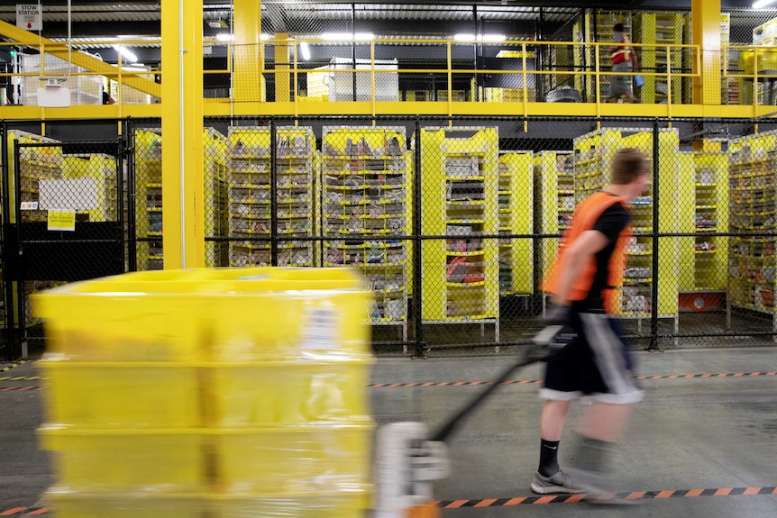 A worker with a high vis vest pulls a pallet jack carrying plastic crates in a warehouse with many shelves and storage units.