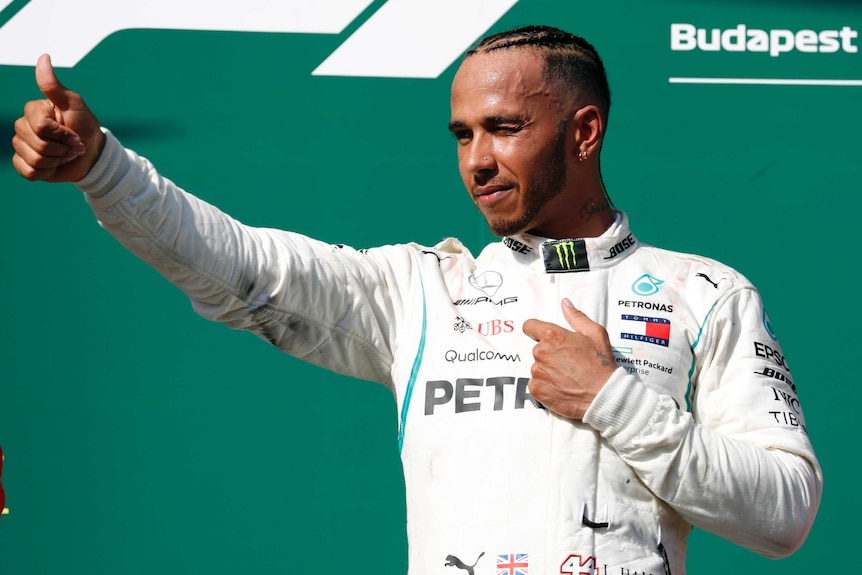 Lewis Hamilton stands on the podium and squints into the sun, giving fans the thumbs up