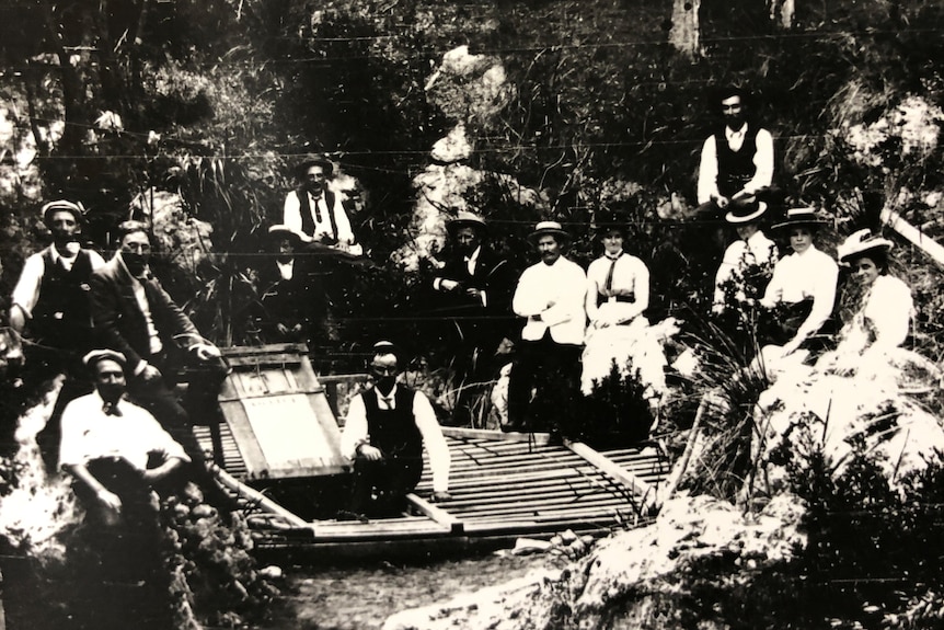 A large group of men and women sitting and standing around a cave entrance in forest. 