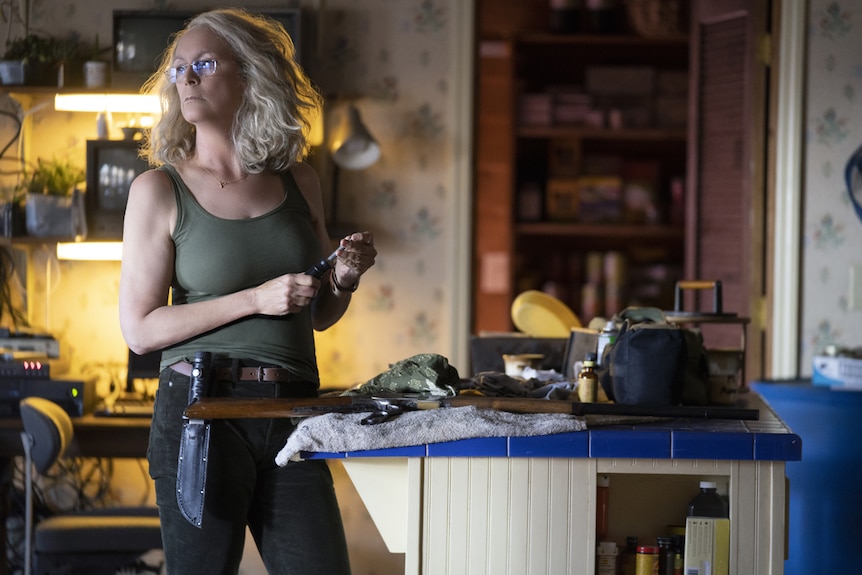 Jamie Lee Curtis with wavy shoulder-length hair in army-green tank top and black jeans, wearing tool belt with massive knife.