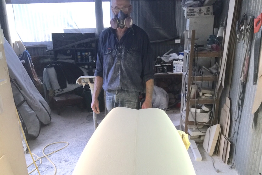 A man wearing a paint mask in a shed with a surfboard in the foreground.