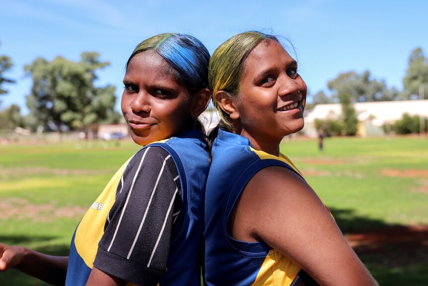 Two young Aboriginal women with died yellow and blue hair stand back to back smiling with football ground in background