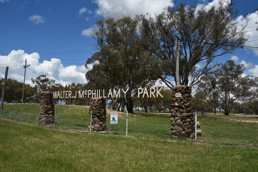 Three cobblestone mounds supporting a sign that says Walter J McPhillamy Park are surrounded by green grass and trees.