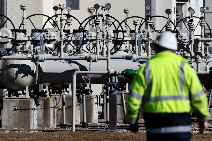 A man in high-vis and a helmet walks in front of pipes.