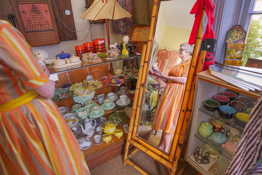 A woman in a yellow and orange dress holds a ceramic doll in the reflection of a standing wooden mirror.