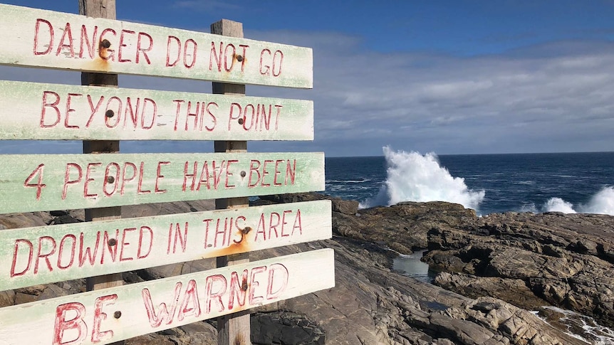A sign warning that other people have died at the location with rocks and waves behind