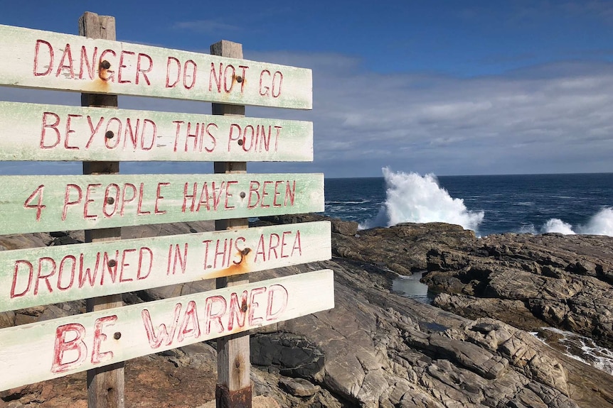 A sign warning that other people have died at the location with rocks and waves behind