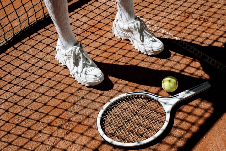 A tennis racquet is seen on the ground of a clay court with a person wearing white shoes on the left.