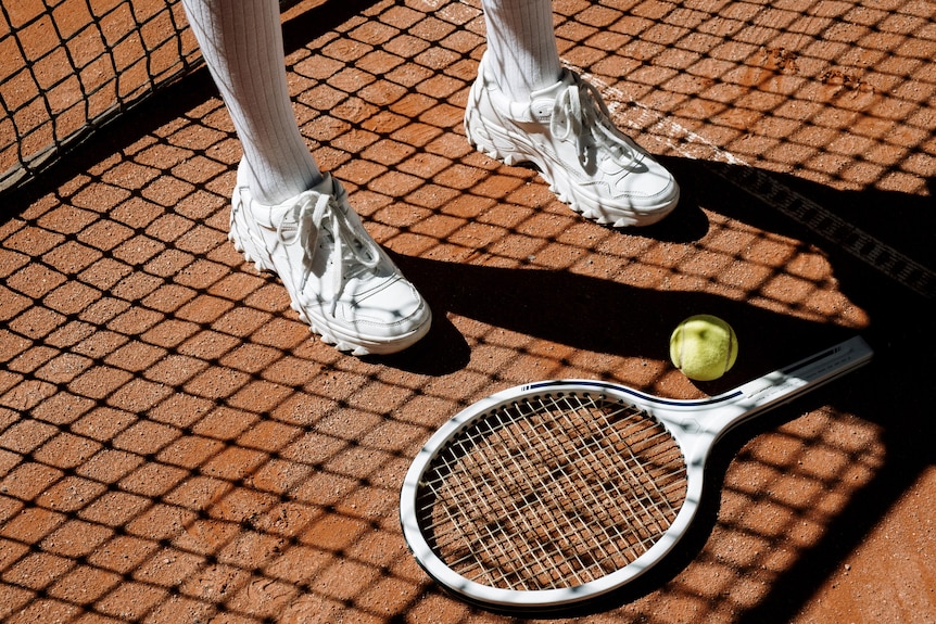 A tennis racquet is seen on the ground of a clay court with a person wearing white shoes on the left.