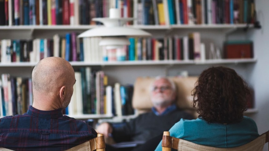 The backs of a man and woman seated separately, with a blurred third person, a man, seated across from the couple, facing them.