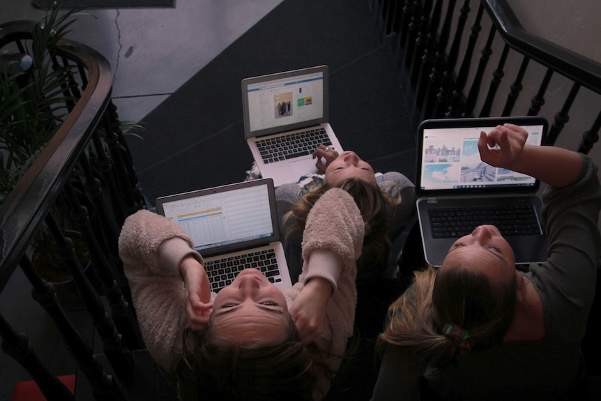 Three women with laptops sit on stairs.