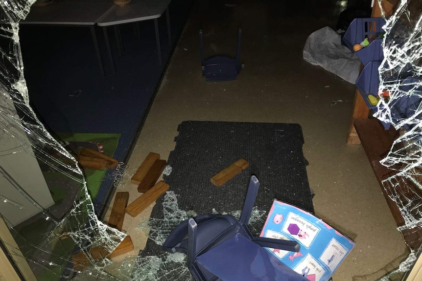 A smashed window with broken glass, pieces of wood and plastic chairs lying on the floor of a classroom.