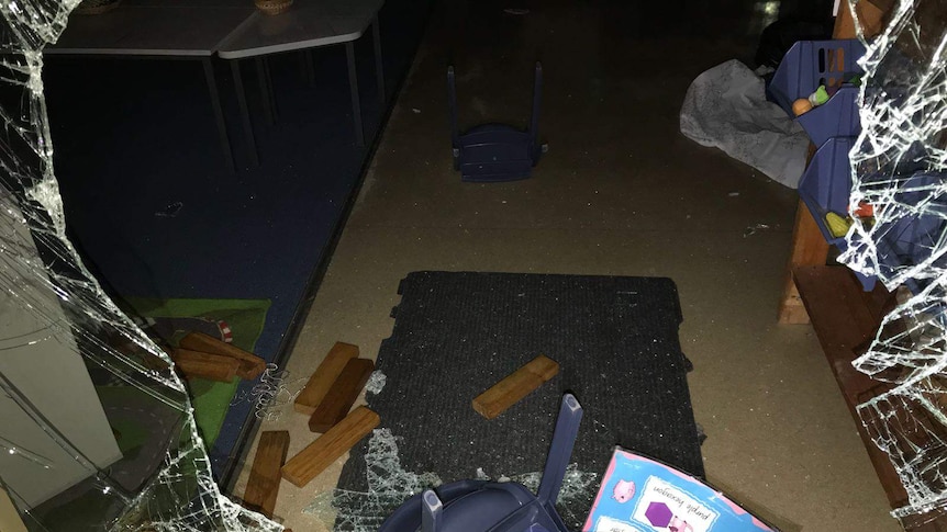 A smashed window with broken glass, pieces of wood and plastic chairs lying on the floor of a classroom.