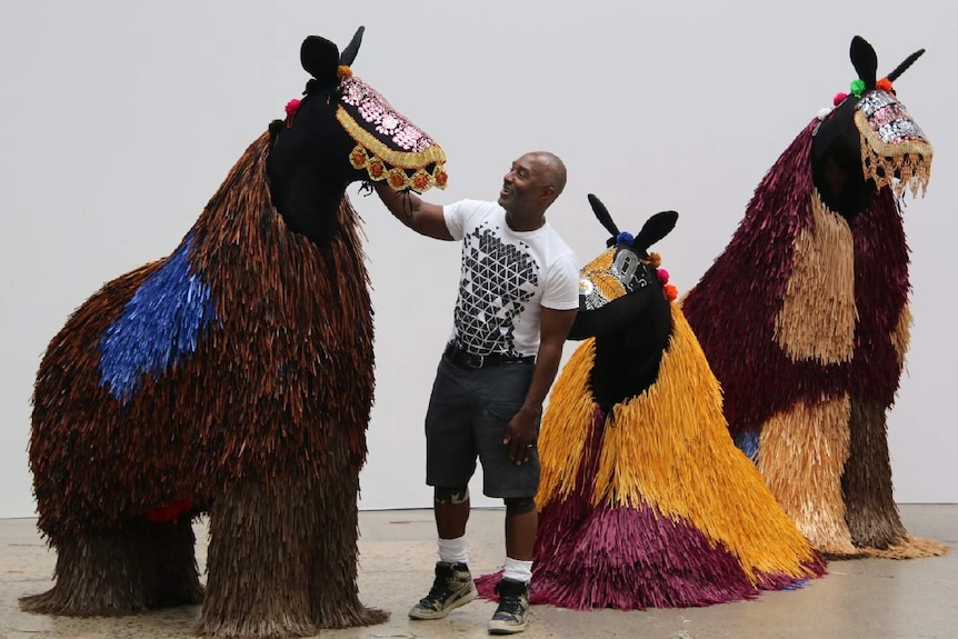 Colour photo of artist Nick Cave smiling and standing with three horse-like sculptures.