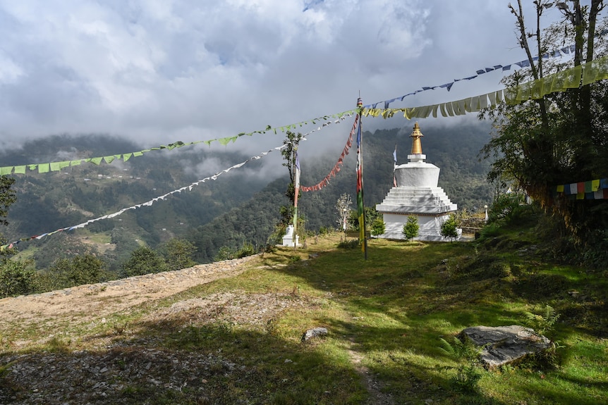 a white stupa on a mountainside surrounded by green grass and forest with mist in the distance 