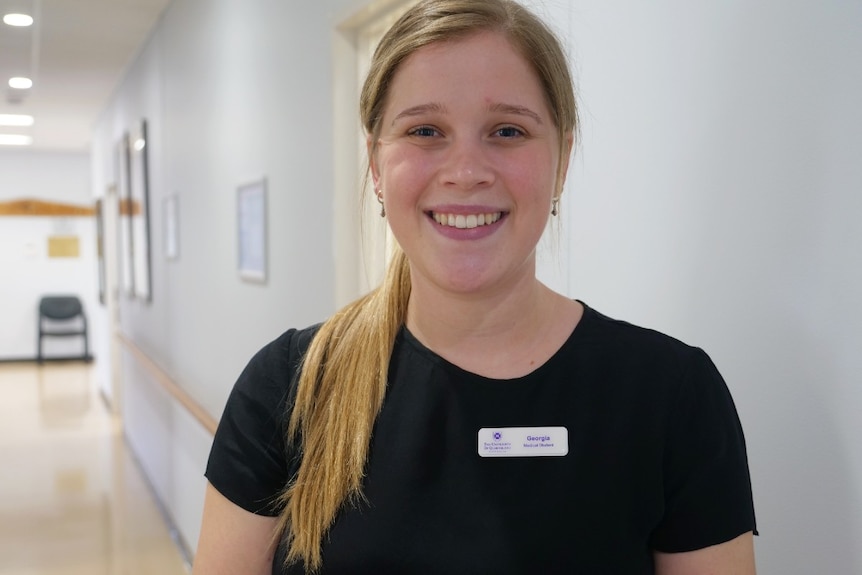 Georgia Austin smiles at the camera, blue eyes, blonde and light brown hair, black shirt, name tag, doctor's surgery behind.