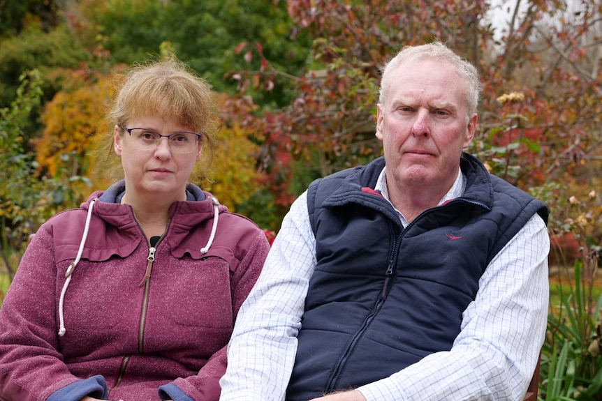 A middle-aged woman and a man sitting outside, dressed for warmth and looking solemn.