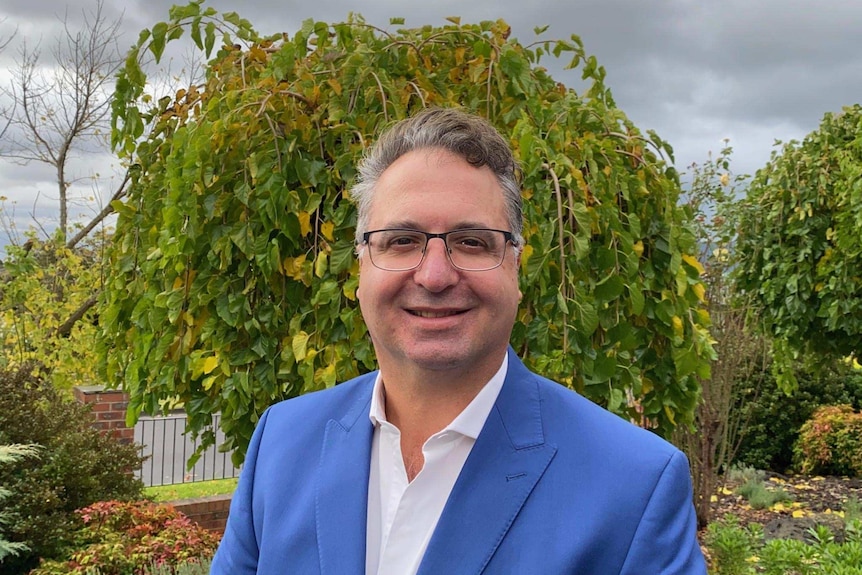 A smiling man stands in a garden wearing a light blue jacket.