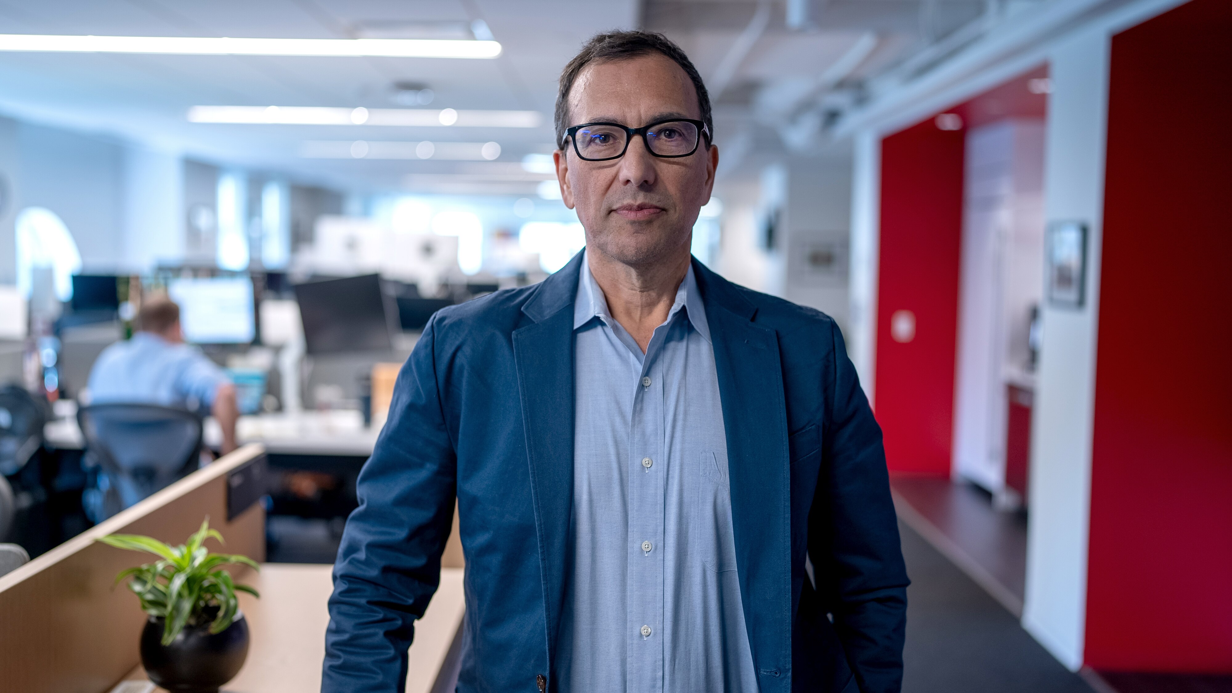 A middle-aged man wearing glasses, a blue suit jacket and blue business shirt standing in an office.