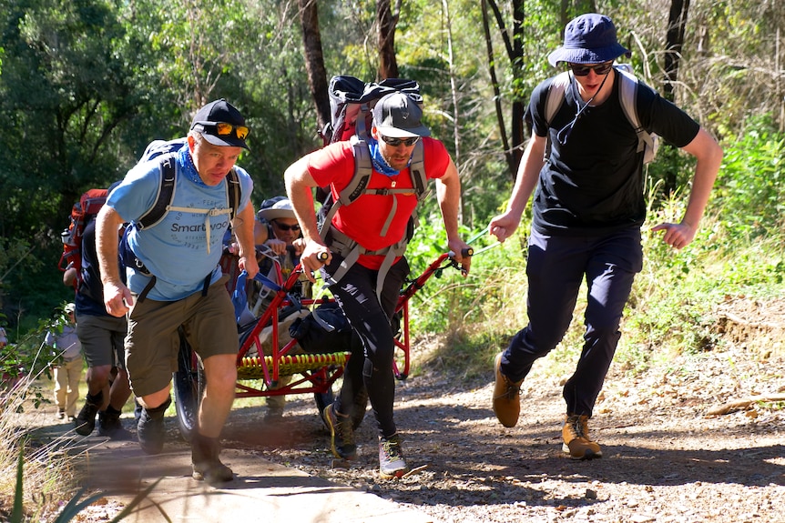 A group of people walk through the countryside, the first two pushing and pulling a cart.