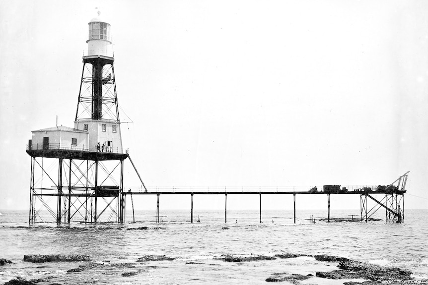 Black and white photo of a large lighthouse connected to a long jetty.