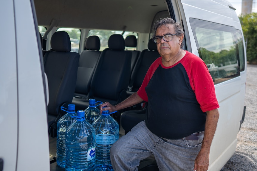 Indigenous man wearing black top with red sleeves and jeans sitting in a van with large bottles of water.