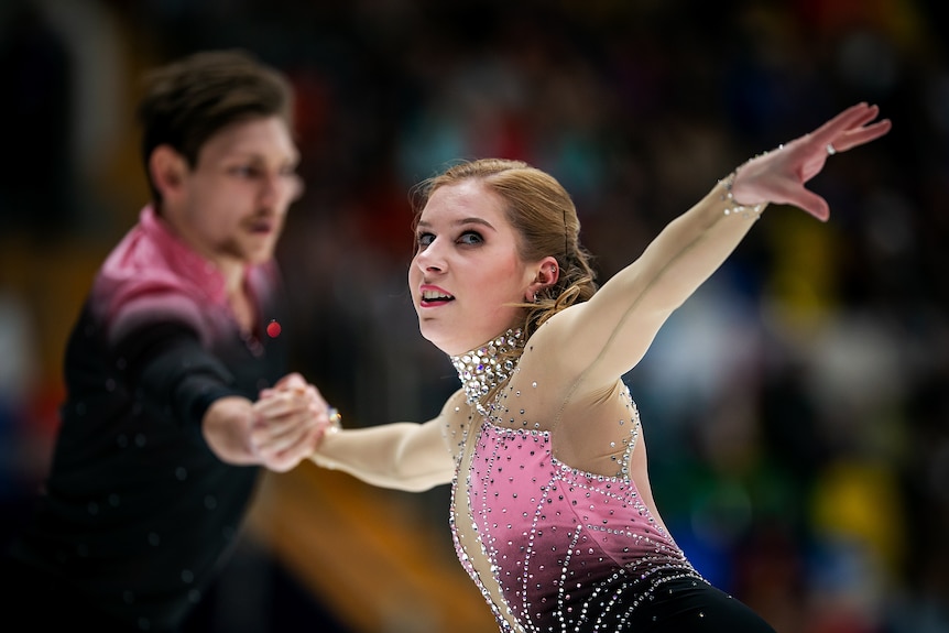A female figure skater looks up holding a pose in a routine as she holds hands with her male partner in the background.