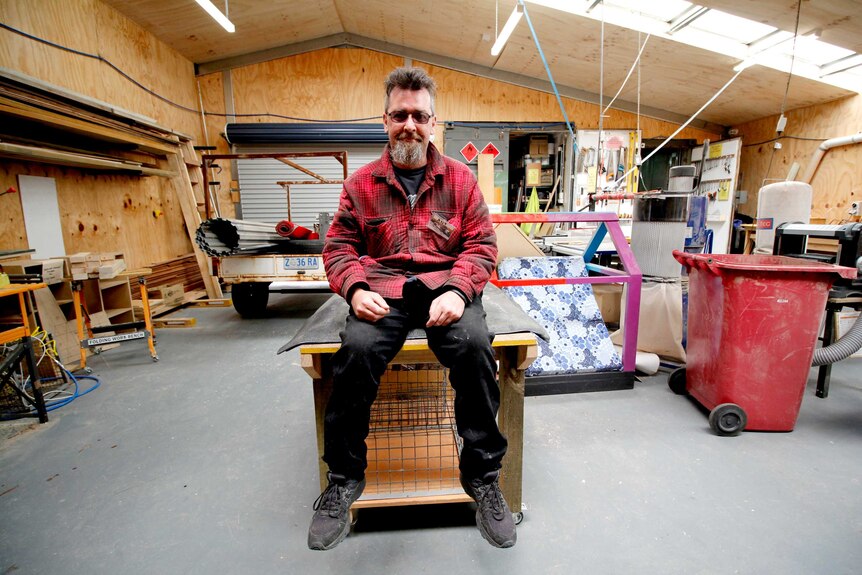 A man sits on a workbench in a men's shed surrounded by equipment