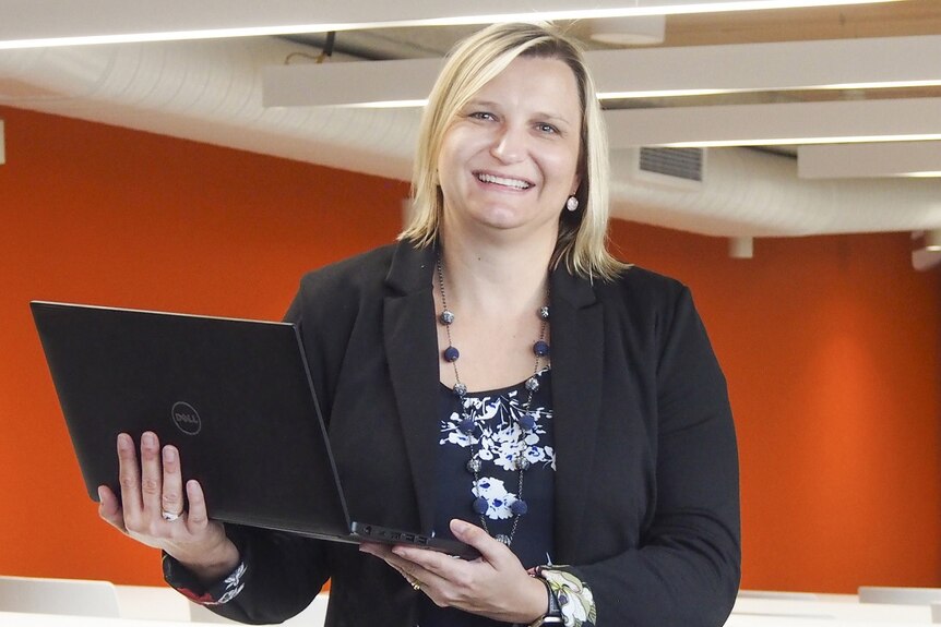 A woman with shoulder-length blonde hair, smiling and holding a laptop with an orange wall in the background.