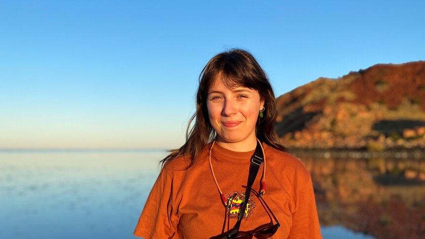 A woman in an orange t shirt stands in front of a calm ocean with red rocks in the distance behind her