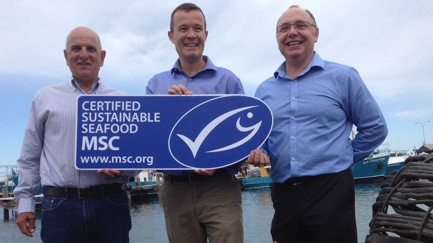 Three men stand at a fishing wharf holding a large cardboard cut out of the certification label, with a bucket of prawns.