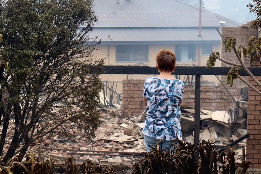 A woman with her back to the camera stands in front of the burnt ruins of a home.