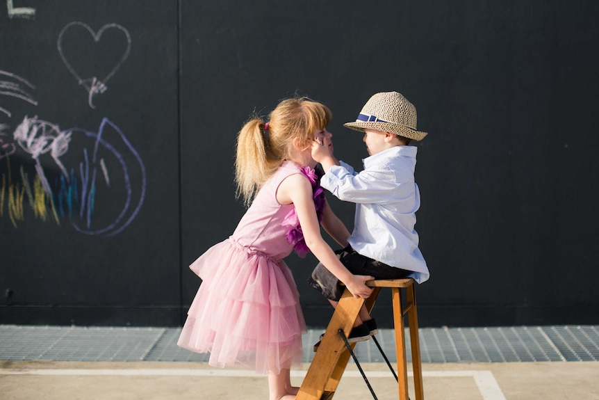 A girl in a pink tutu looks into her little brother's eyes while he affectionally pinches her facial cheeks.