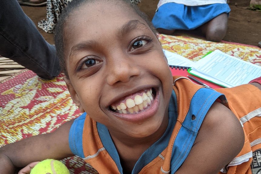 A young child with big beautiful eyes, who has cerebral palsy, lays on a mat and holds a tennis ball smiling widely 