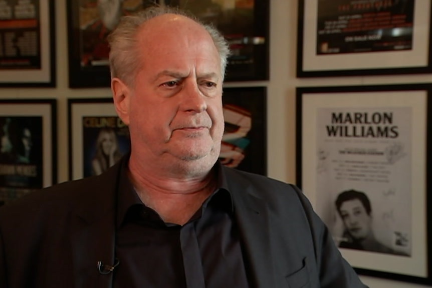 A man in a black suit standing in front of a wall featuring framed music posters.