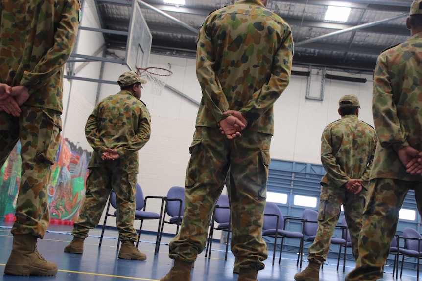 Five teenage boys in camouflage army uniforms face away from the camera with their hands behind their backs.