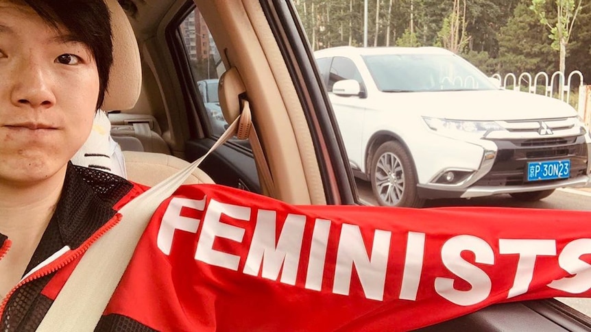 A woman in a car with the word 'feminist' written on the sleeve of her jacket