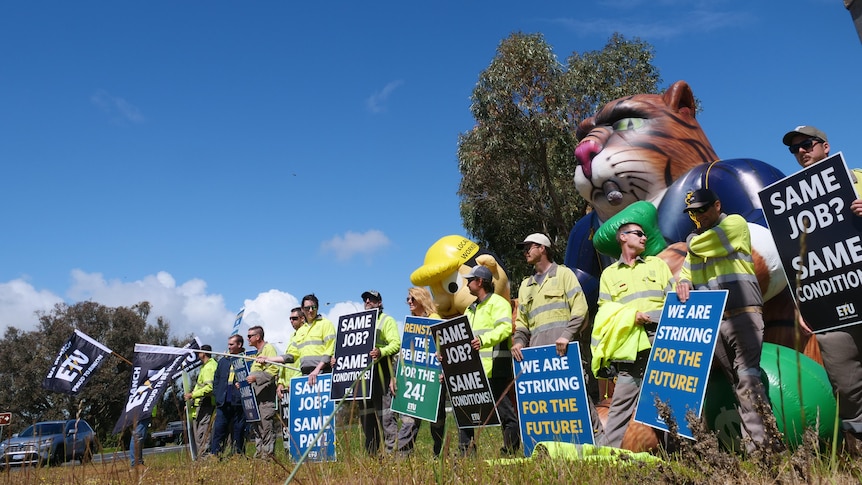 Workers holding protest signs and union flags during a strike, with a giant inflatable cat in the background.