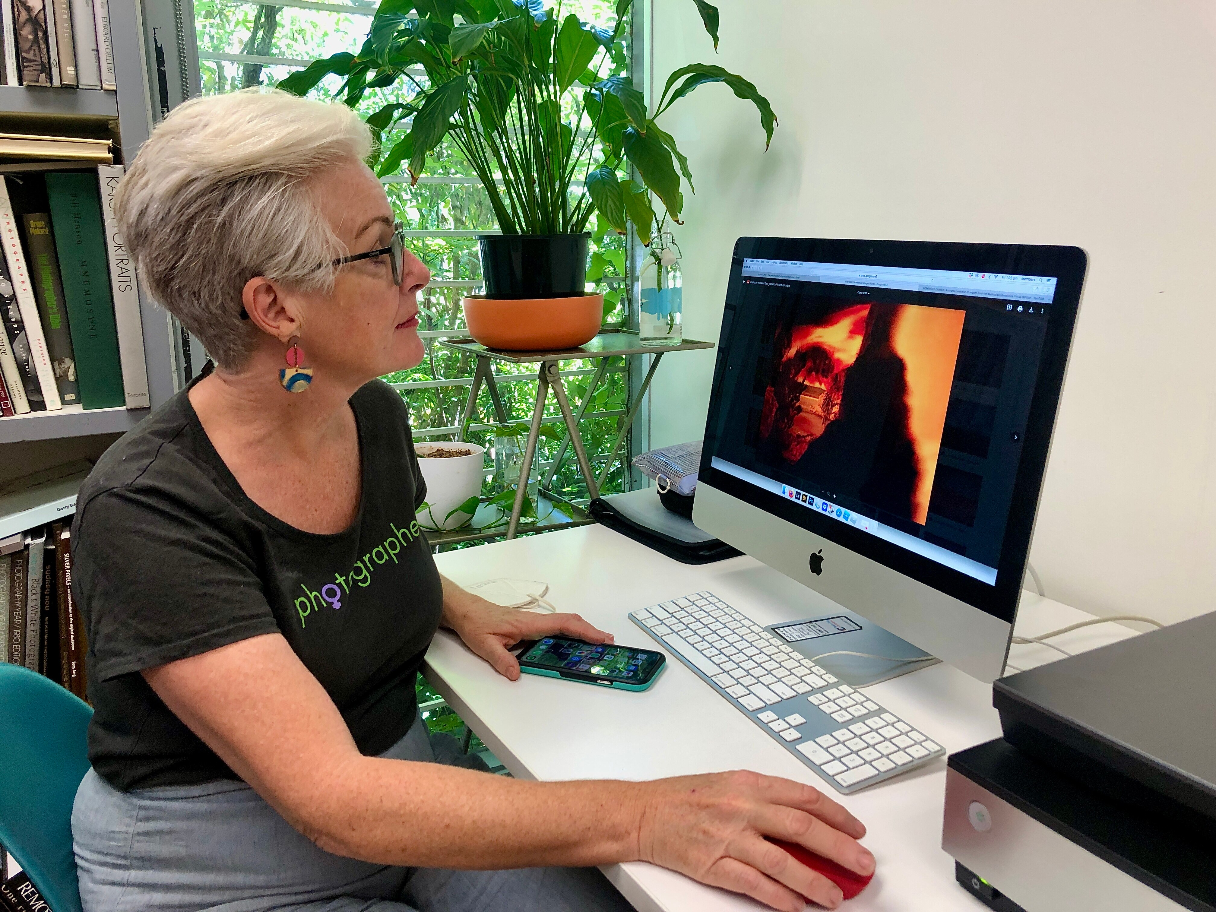 A woman with short grey hair and glasses looks at a photograph of a fire on a computer screen.