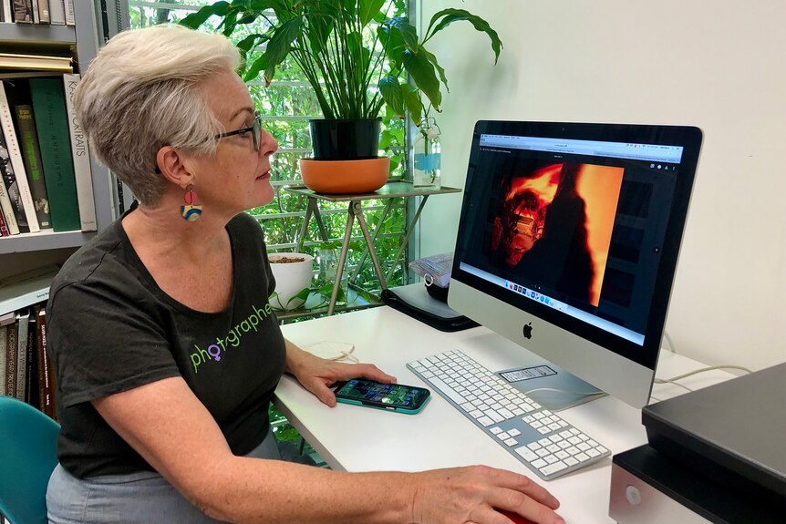 Une femme aux cheveux gris courts et à lunettes regarde une photo d'un incendie sur un écran d'ordinateur.