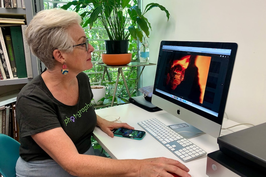 A woman with short grey hair and glasses looks at a photograph of a fire on a computer screen.