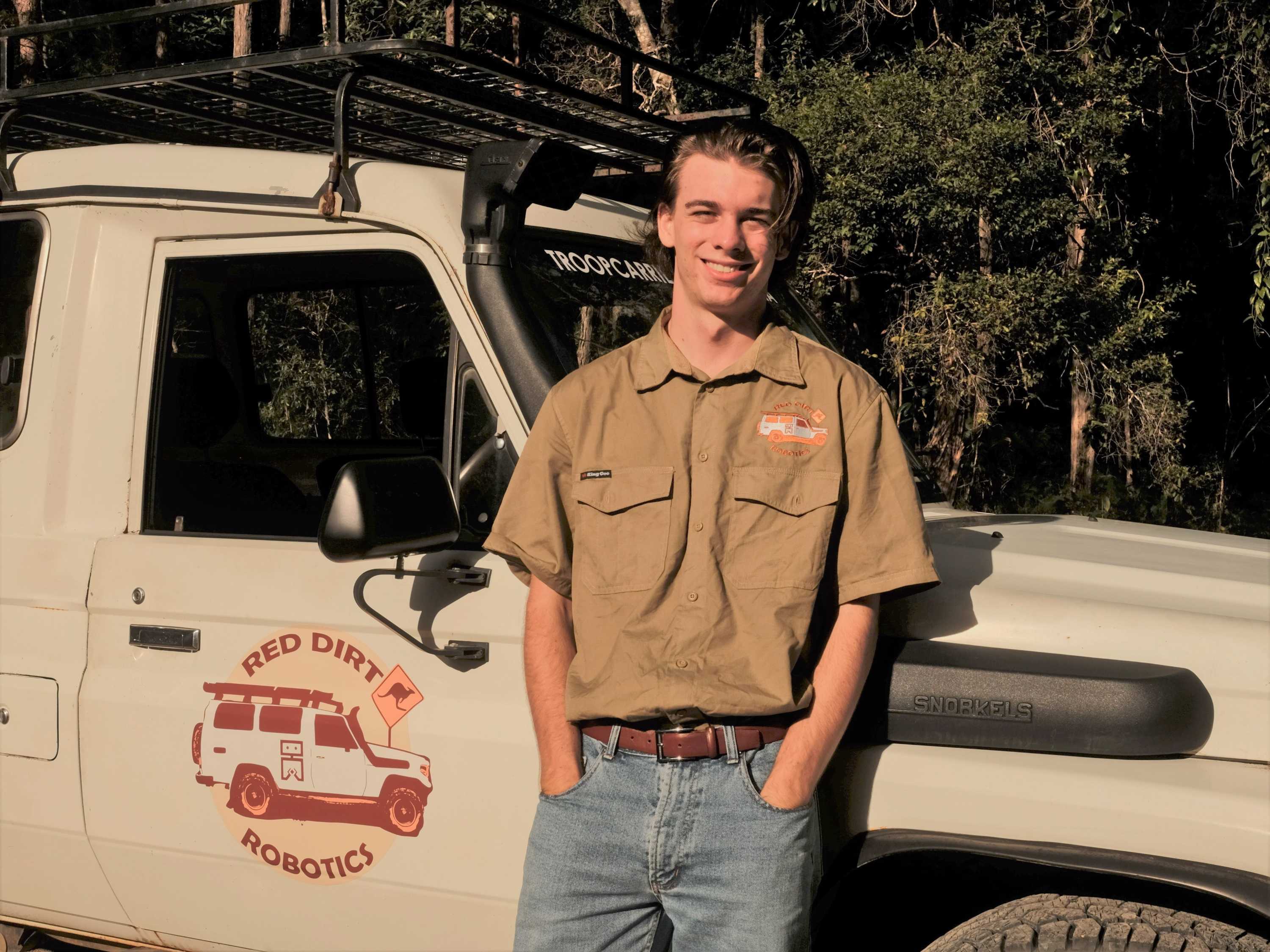 Jameson stands in front of his 4wd smiling to the camera wearing a brown shirt with a logo saying Red Dirt Robotics