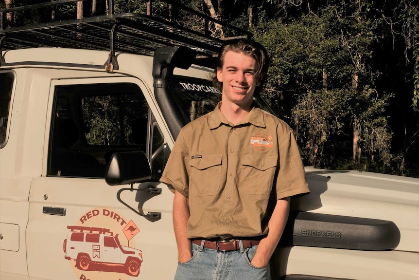 Jameson stands in front of his 4wd smiling to the camera wearing a brown shirt with a logo saying Red Dirt Robotics
