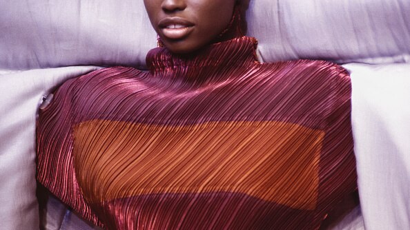 A close up of a model's chest shows a vibrant pleated top with a high neck in autumnal tones.